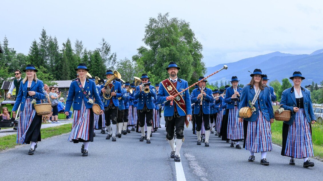 Eine Musikkapelle in traditioneller Tracht marschiert auf der Straße. Im Hintergrund sind Bäume und Berge zu sehen. | © Michael Simonlehner