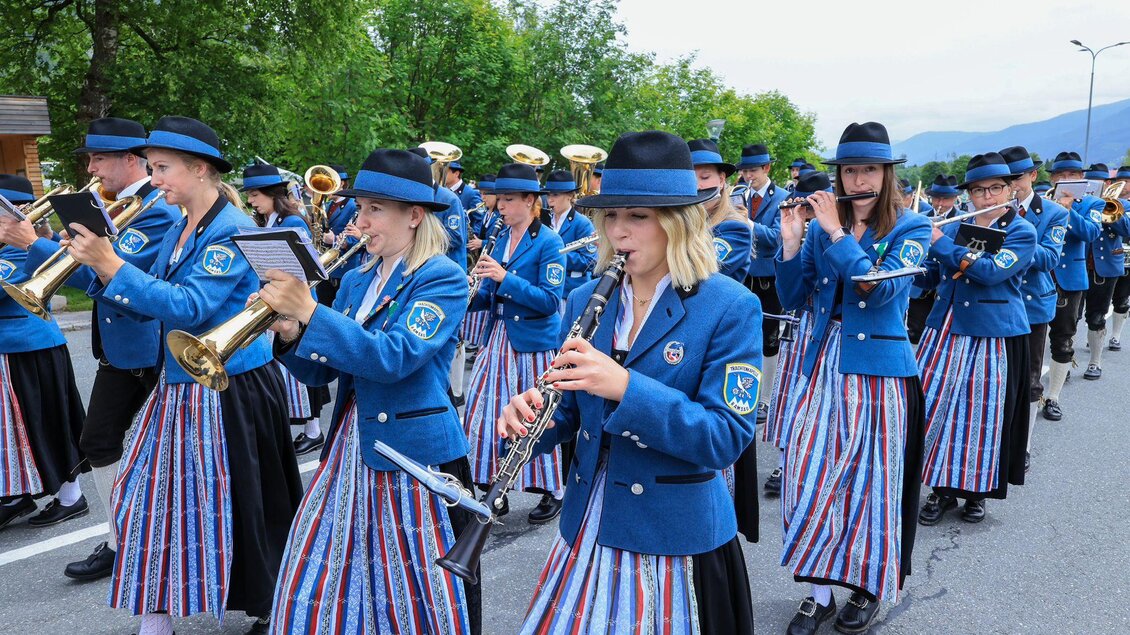 Eine musikalische Parade mit Musikerinnen und Musikern in blauen Uniformen und gestreiften Röcken. Sie spielen verschiedene Blasinstrumente und gehen eine Straße entlang. | © Michael Simonlehner
