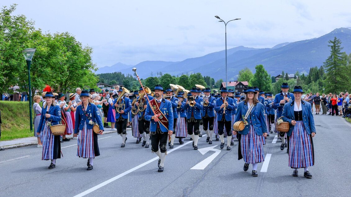 Ein farbenfroher Umzug mit Menschen in traditionellen Trachten. Die Teilnehmer tragen Musikinstrumente und gehen durch eine ruhige Straße, umgeben von Bäumen und Bergen. | © Michael Simonlehner