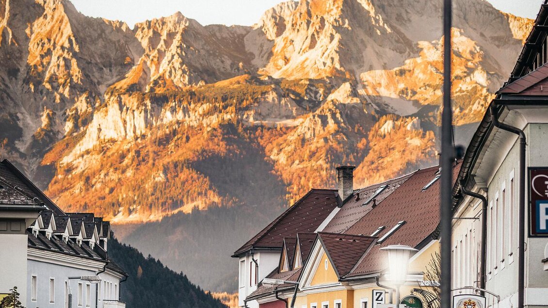 Eine ruhige Straße in einer kleinen Stadt mit bunten Gebäuden und einem beeindruckenden Bergpanorama im Hintergrund. Die Sonne beleuchtet die Berge und verleiht der Szene eine warme Atmosphäre. | © TV Gesäuse