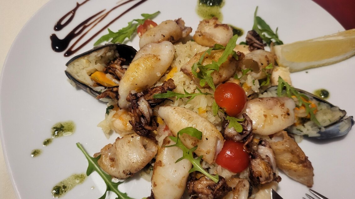 Ein Teller mit gegrilltem Meeresfrüchte, einschließlich Tintenfisch und Muscheln, garniert mit frischen Kräutern und Kirschtomaten. Dazu liegt ein Stück Zitrone und ein Balsamico-Dressing. | © Trattoria la Fortuna