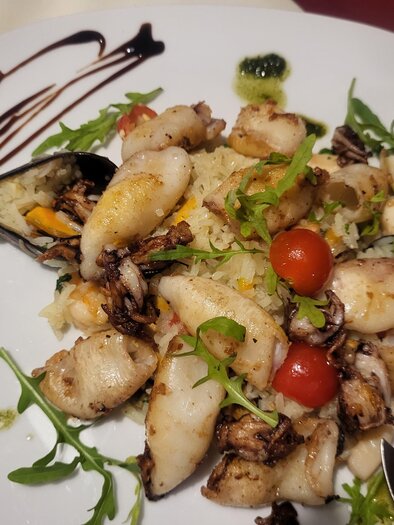 Ein Teller mit gegrilltem Meeresfrüchte, einschließlich Tintenfisch und Muscheln, garniert mit frischen Kräutern und Kirschtomaten. Dazu liegt ein Stück Zitrone und ein Balsamico-Dressing. | © Trattoria la Fortuna