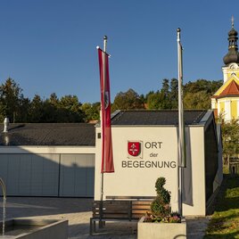 Ein modernes Gebäude mit der Aufschrift "Ort der Begegnung" steht vor einer historischen Kirche. Umgeben von Bäumen und einem klaren Himmel. | © Kurkommission Bad Blumau