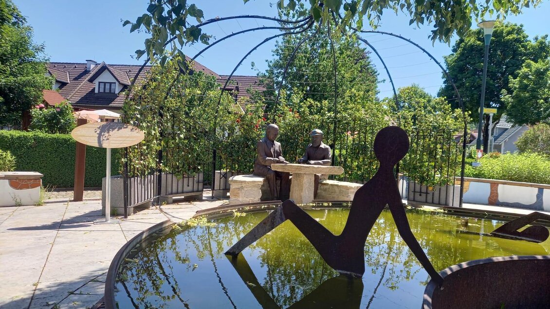 Ein schöner Garten mit einer Skulptur und einem Teich. Zwei Menschenfiguren sitzen an einem Tisch unter Bäumen. | © Thermen- & Vulkanland