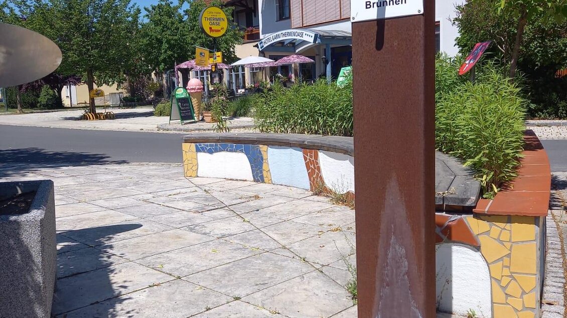 Ein Trinkbrunnen mit einem Schild an einer Straße. Im Hintergrund sind mehrere Gebäude und Bäume sichtbar. | © Thermen- & Vulkanland