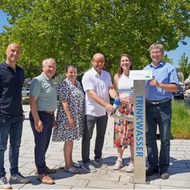 Eine Gruppe von fünf Personen steht um einen Trinkwasserbrunnen. Sie lächeln und posieren in einer städtischen Umgebung mit Bäumen im Hintergrund. | © die Neue Stadt Feldbach