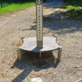 Ein Trinkbrunnen auf einem Weg, umgeben von grüner Landschaft. Der Brunnen steht auf einem kleinen, schottrigen Platz. | © Marktgemeinde Burgau