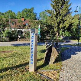 Ein Trinkbrunnen steht auf einer Wiese, umgeben von Bäumen und einer Parkbank. Im Hintergrund sieht man ein Wohngebäude und Straßenlaternen. | © Thermen- & Vulkanland