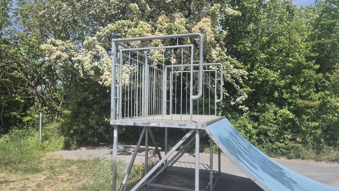 Eine Spielplatzkonstruktion mit einer Rutsche und einem erhöhten Spielturm ist vor einem grünen Hintergrund zu sehen. Die Umgebung ist von Bäumen und Sträuchern umgeben. | © Petra Stossier