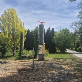 Ein Weg führt durch eine grüne Landschaft mit Bäumen und Sträuchern. Im Vordergrund steht ein hoher Pfosten mit einer farbigen Markierung. | © Petra Stossier