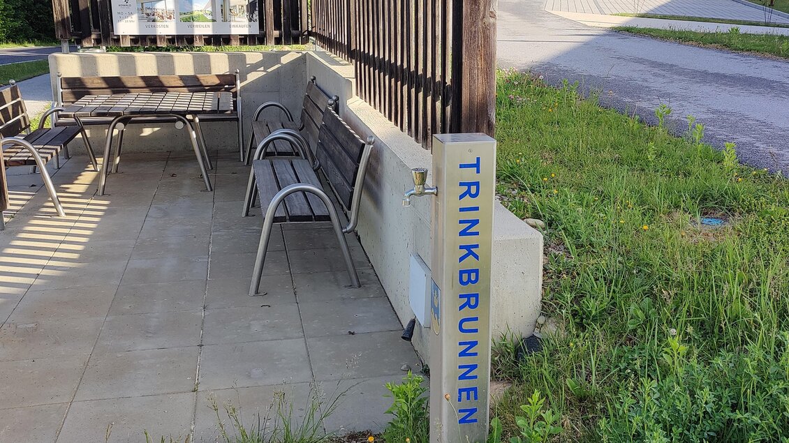 Ein Trinkbrunnen steht an einem kleinen Sitzbereich mit Stühlen. Im Hintergrund sind einige Häuser und eine Straße zu sehen. | © Thermen- & Vulkanland