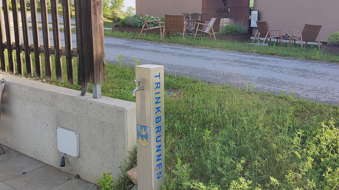 Ein Trinkbrunnen steht an einem Weg, umgeben von Grünflächen. Im Hintergrund sind einige Gartenmöbel und ein modernes Gebäude zu sehen. | © Thermen- & Vulkanland