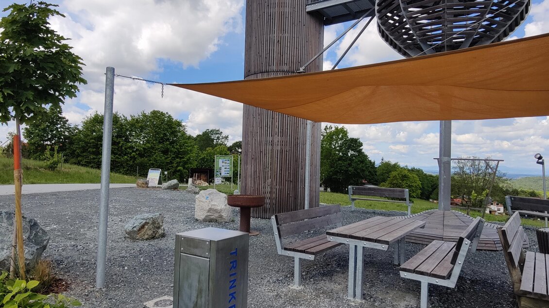 Eine moderne Aussichtsplattform mit einem hohen, schornsteinartigen Turm und einem spiraligen Geländer. Der Bereich ist mit Sitzgelegenheiten und Sonnensegeln ausgestattet, um einen schönen Ausblick zu genießen. | © Thermen- & Vulkanland