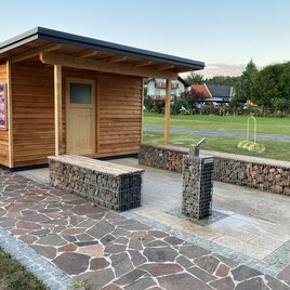 Ein kleiner Holzschuppen steht auf einem gepflasterten Platz. Daneben befindet sich eine Sitzgelegenheit und eine Wassersäule in einem gepflegten Garten. | © Gemeinde Unterlamm