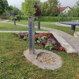 Ein Trinkbrunnen steht in einem gepflegten Park mit bunten Blumenbeeten. Der Weg rund um den Brunnen ist aus Steinen und von Grünflächen umgeben. | © Thermen- & Vulkanland