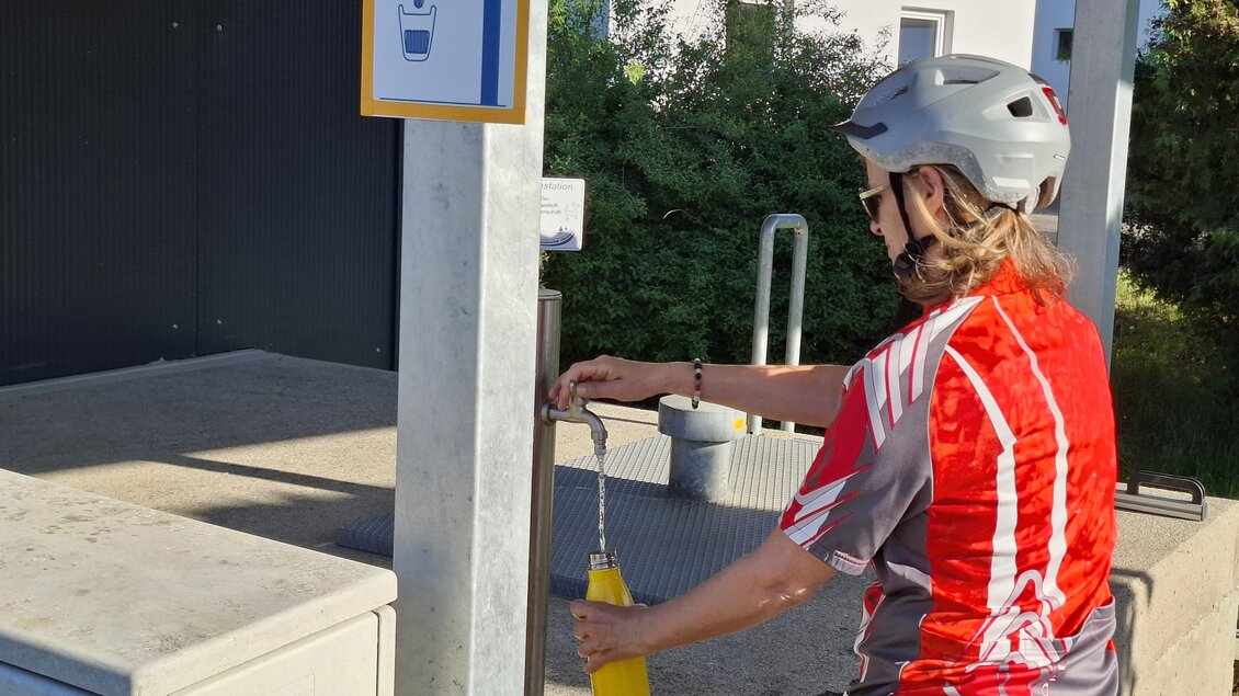 Eine Person mit einem Fahrradhelm füllt eine Wasserflasche an einem Trinkbrunnen auf. Im Hintergrund sind Bäume und eine Trainingsanlage zu sehen.