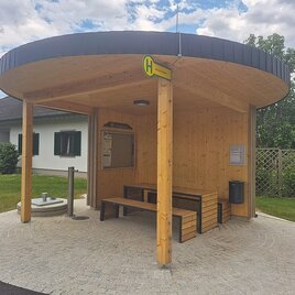 Eine moderne Bushaltestelle aus Holz mit einem runden Dach. Der Bereich ist sauber und einladend, umgeben von Grünflächen. | © Petra Stossier