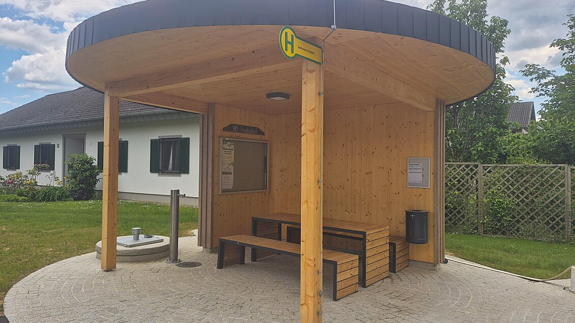 Eine moderne Bushaltestelle aus Holz mit einem runden Dach. Der Bereich ist sauber und einladend, umgeben von Grünflächen. | © Petra Stossier