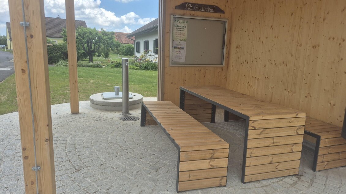Ein überdachter Sitzbereich mit Holzbänken und einem Tisch. Im Hintergrund sind Grünflächen und ein klarer Himmel zu sehen. | © Petra Stossier