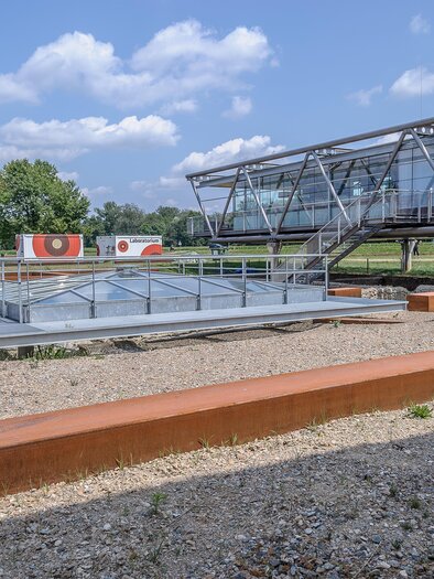 A modern building with large windows and a covered area stands next to a gravel garden. In the background, there are trees and a blue sky. | © Universalmuseum um Joanneum N. Lackner