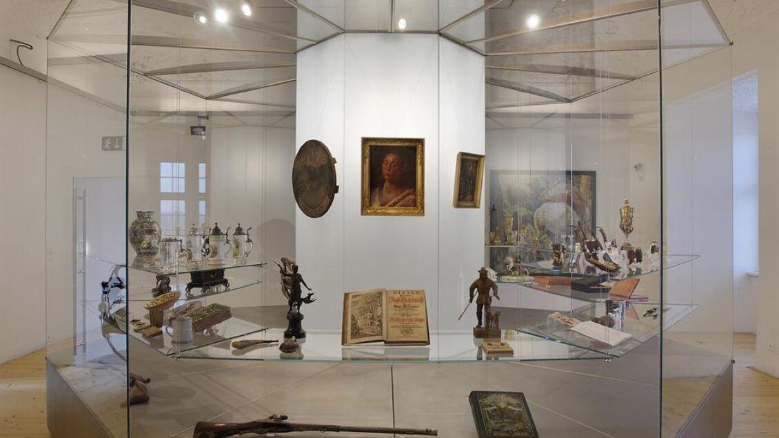 Ein gläserner Ausstellungsstand mit verschiedenen Kunstgegenständen und Antiquitäten. Im Hintergrund sind Gemälde und Skulpturen zu sehen. | © Universalmuseum Joanneum - M. Wimmler