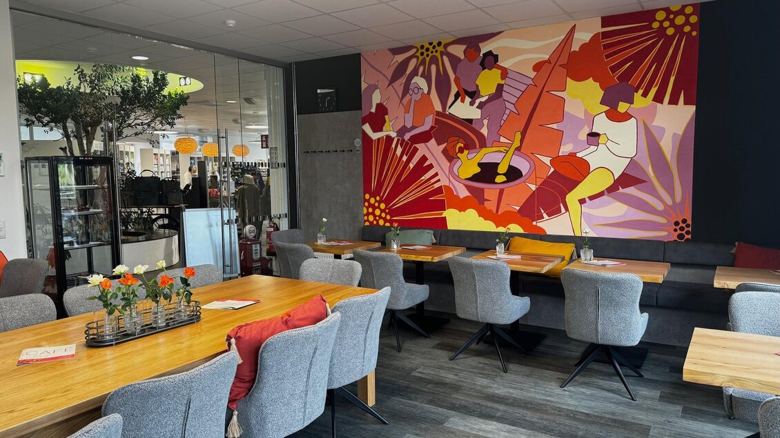 Ein modernes Café mit eleganten grauen Stühlen und Holztischen. An der Wand hängt ein buntes Kunstwerk, das eine lebendige Szene darstellt. | © Unser Cafe'