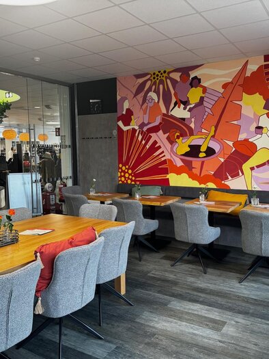Ein modernes Café mit eleganten grauen Stühlen und Holztischen. An der Wand hängt ein buntes Kunstwerk, das eine lebendige Szene darstellt. | © Unser Cafe'