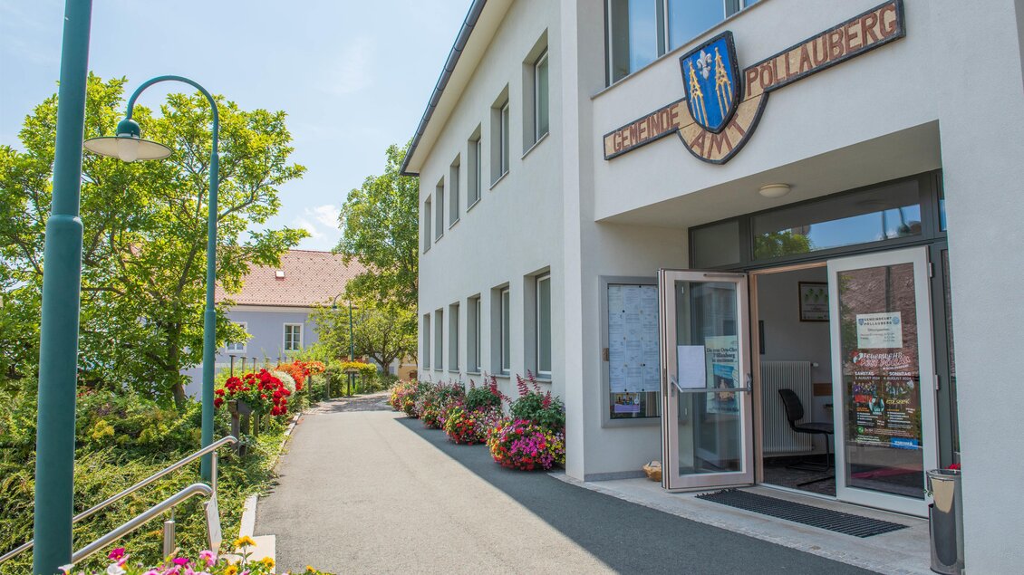 Ein modernes Gebäude mit dem Schild „Gemeinde Pohlheim“. Der Eingang ist von blühenden Pflanzen und Bäumen umgeben. | © Helmut Schweighofer