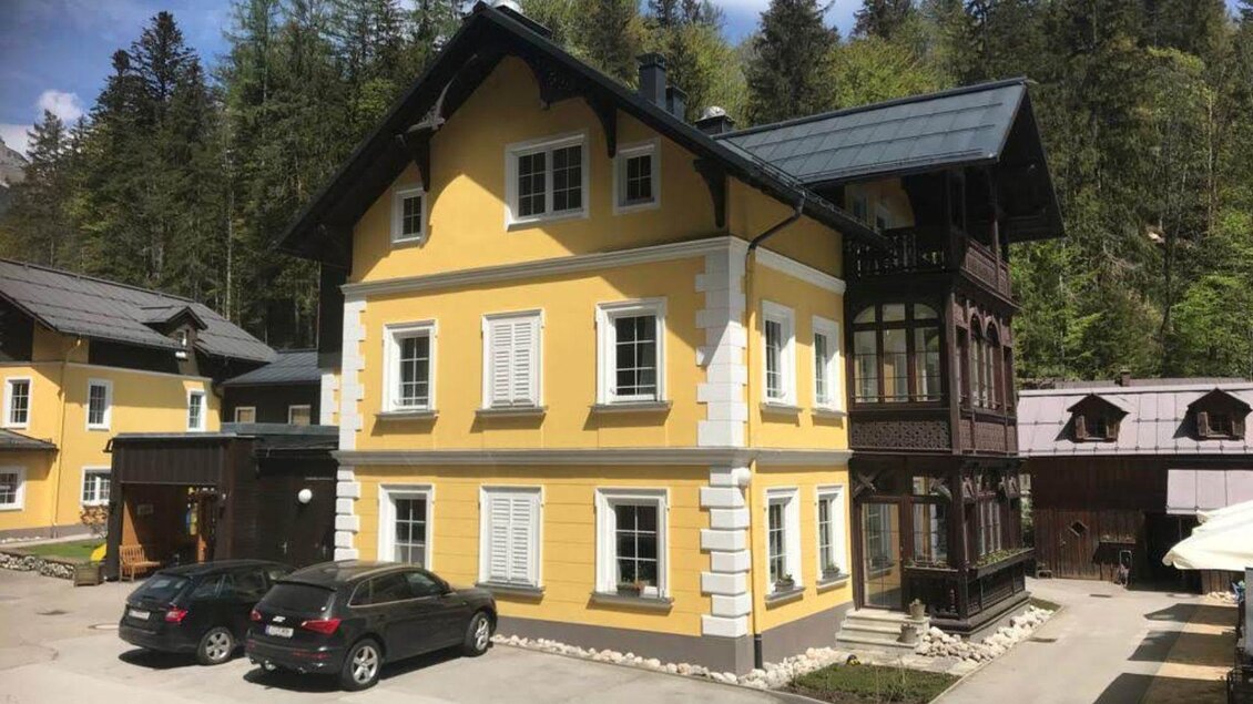 Das schön gelb gestrichene Gebäude mit Holzverkleidung und Fensterläden ist die Villa Styria, ein Hotel in Bad Aussee. Es steht in einer grünen Umgebung mit Bäumen im Hintergrund. | ©  Villa Styria, Bad Aussee