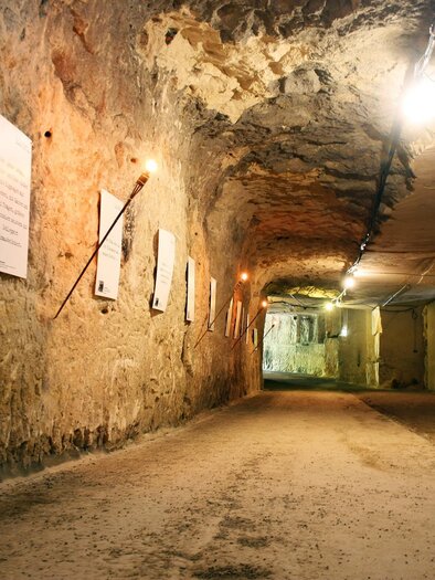 Ein unterirdischer Tunnel mit beleuchtetem Fels und Informationsplakaten an den Wänden. Der Boden ist aus Erde und der Raum wirkt geräumig und historisch. | © Stefan Matic