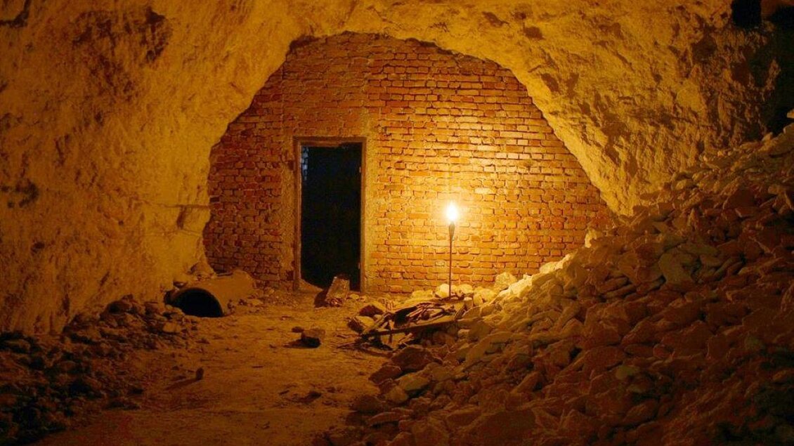 Ein dunkler Tunnel mit einer gemauerten Wand und einer Tür am Ende. Eine einzelne Lampe beleuchtet den Raum und wirft Schatten auf den Boden. | © Stefan Matic