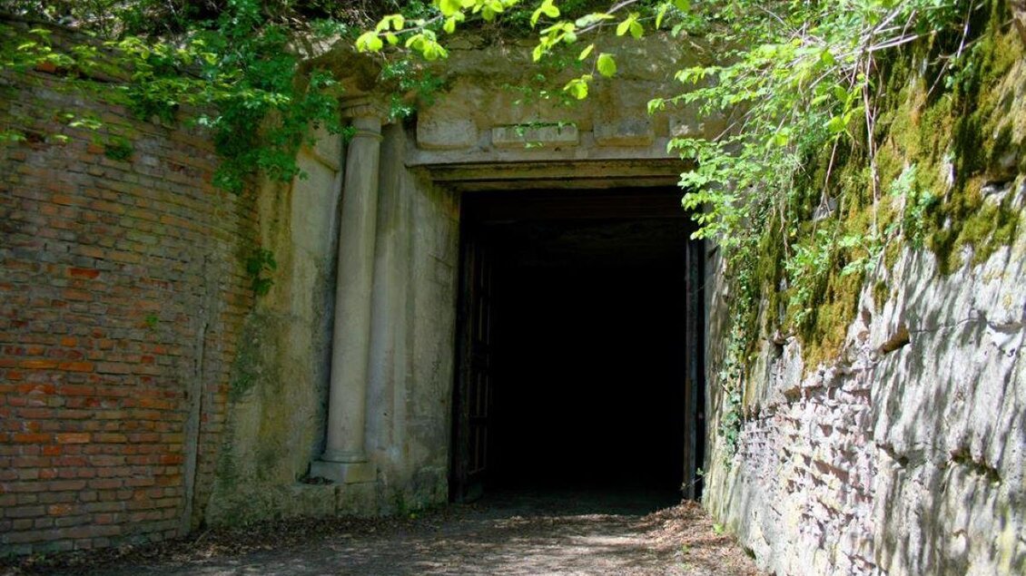 Ein dunkler Eingang in eine Höhle, umgeben von grünen Bäumen und moosbedeckten Felsen. Die Wände sind aus Ziegeln und Stein, was eine alte, verlassene Atmosphäre schafft. | © Stefan Matic