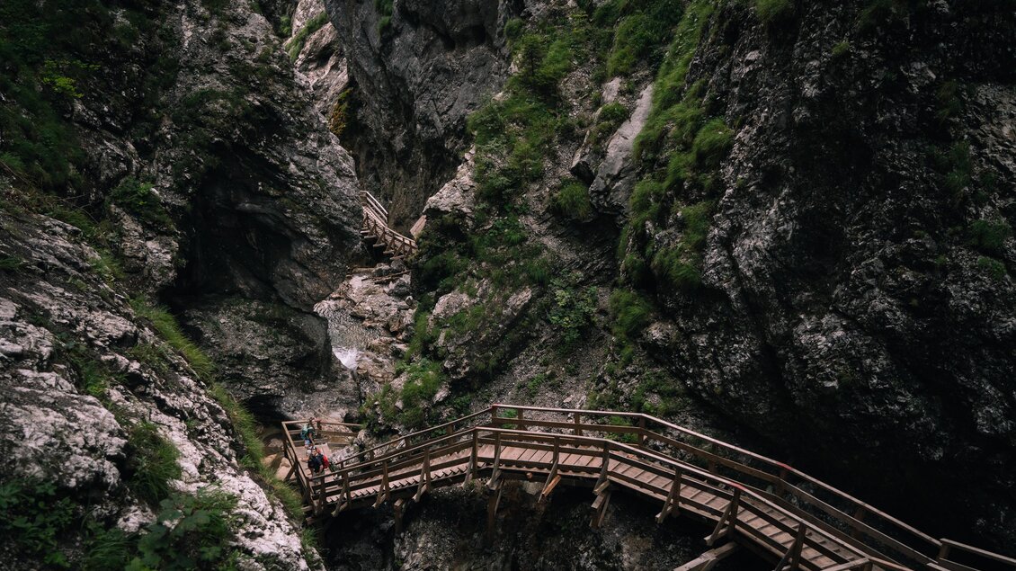 Das Bild zeigt eine enge, steile Felsenschlucht mit etwas Grünbewuchs. Ein hölzerner Steg mit Treppen und Geländern führt hindurch. Zwei Wandernde mit Rucksäcken sind darauf unterwegs, unter ihnen fließt ein schmaler Bach. | © Christoph Lukas