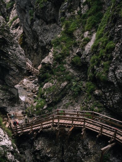 Das Bild zeigt eine enge, steile Felsenschlucht mit etwas Grünbewuchs. Ein hölzerner Steg mit Treppen und Geländern führt hindurch. Zwei Wandernde mit Rucksäcken sind darauf unterwegs, unter ihnen fließt ein schmaler Bach. | © Christoph Lukas