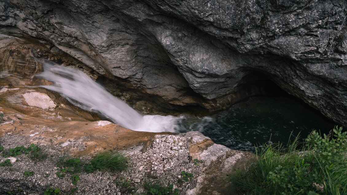 Das Bild zeigt einen kleinen Wasserfall, der über glatten Fels in ein dunkles, rundes Becken in einer Felsenhöhle fließt. Umgeben ist die Szene von grauem Gestein und etwas grünem Bewuchs am Rand. | © Christoph Lukas