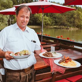 A man stands at a table overlooking a lake, holding two plates with different dishes. The table is surrounded by red umbrellas and flowers. | © Mayr