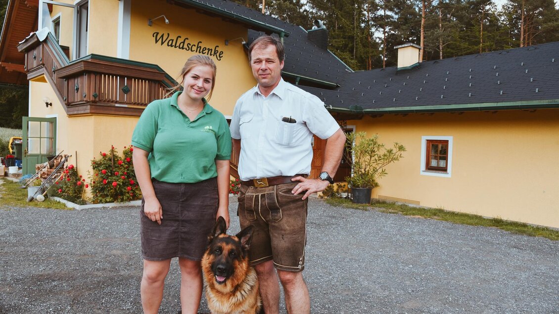 Ein Paar steht vor einem gelben Gebäude mit der Aufschrift "Waldschenke". Neben ihnen sitzt ein Schäferhund. | © Lederer