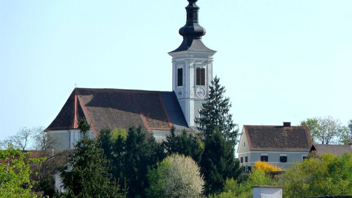 Eine malerische Kirche mit einem hohen Turm, umgeben von Bäumen und grüner Landschaft. Der Himmel ist klar und blau. | © Romana Maier