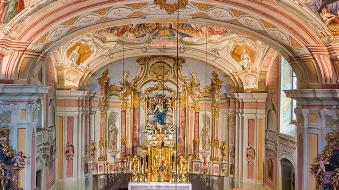 Ein wunderschöner Kirchenraum mit prächtigen Deckenfresken und kunstvollen Verzierungen. Der Altar ist zentral platziert und von Holzbankreihen umgeben. | © Romana Maier