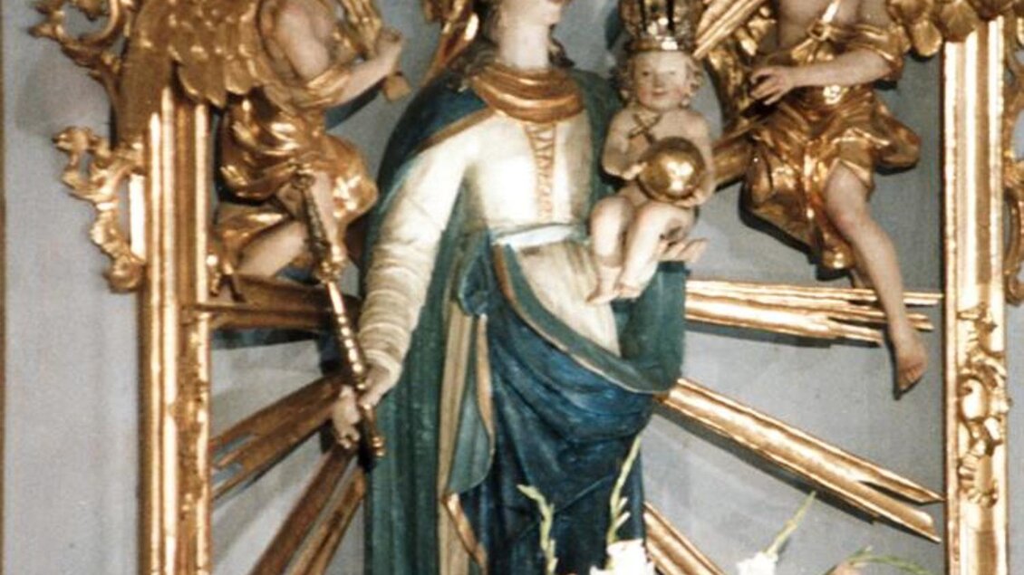 Eine religiöse Statue der Jungfrau Maria, umgeben von Engeln und mit einem Kind in den Armen. Der Hintergrund zeigt dekorative goldene und blaue Elemente. | © Romana Maier