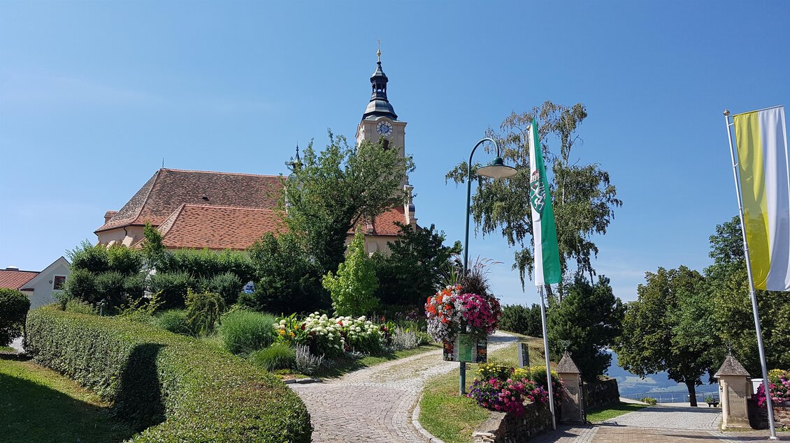 Eine malerische Kirche steht auf einem Hügel, umgeben von grünen Bäumen und bunten Blumen. Der klare Himmel taucht die Szene in helles Licht. | © Tourismusverband Oststeiermark