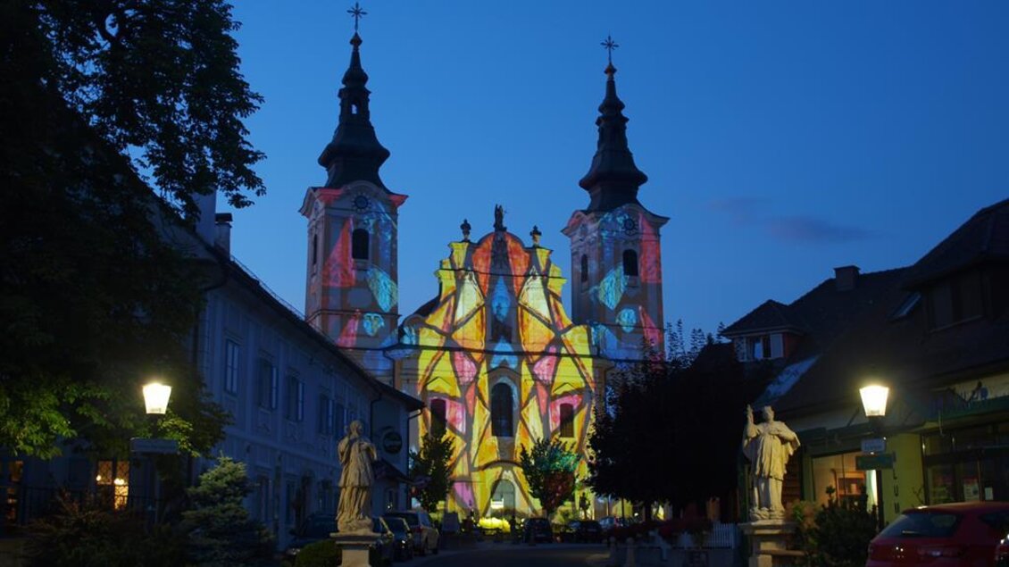 Eine beeindruckende Kirche mit leuchtenden Farben, die die Fassade schmücken. Der Abendhimmel ist blau, und die Umgebung ist ruhig mit Straßenlaternen und Gebäuden. | © Andreas Ruckenstuhl