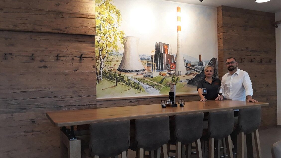Die Gastgeber im Walter's Genusswirt in Voitsberg stehen an der Bar vor einer beeindruckenden Wandmalerei. Das Bild zeigt eine stilvolle Einrichtung mit einer einladenden Holztheke und einem einzigartigen Kunstwerk im Hintergrund. | © TV Lipizzanerheimat