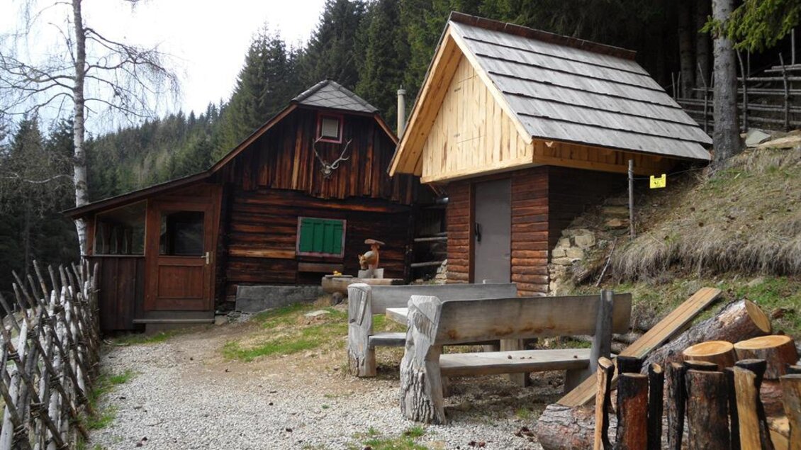 Eine rustikale Hütte aus Holz in einer grünen Waldlandschaft. Daneben steht ein kleiner Anbau und Holzstämme sind gestapelt. | © TV Lipizzanerheimat_EU