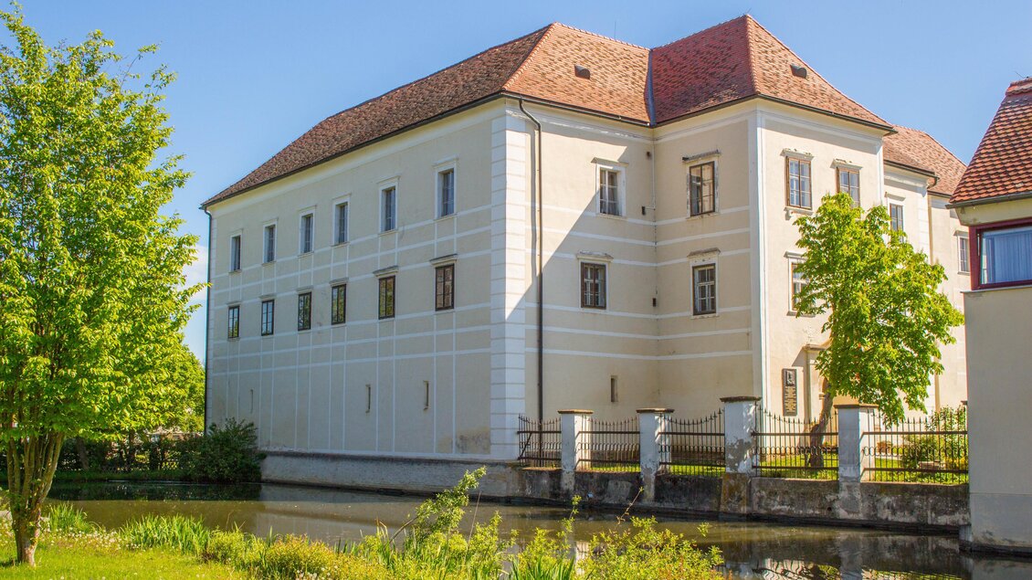 Ein historisches Gebäude mit weißer Fassade und einem roten Dach. Es steht neben einem Teich und ist von grünen Bäumen umgeben. | © Sandra Brünner