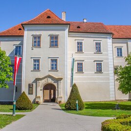 Ein historisches Gebäude mit roten Dächern und großen Fenstern. Der Eingang ist von gepflegten Bäumen und Sträuchern umgeben. | © Sandra Brünner