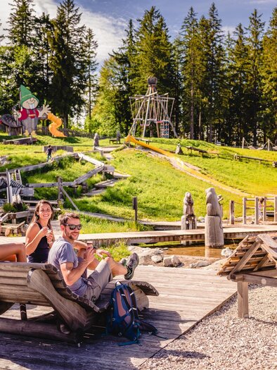 Eine Gruppe von vier Kindern sitzt auf einer Bank und genießt die Zeit im Freien. Im Hintergrund sieht man einen Spielplatz mit vielen Aktivitäten und Bäumen. | © Mariazeller Bürgeralpe