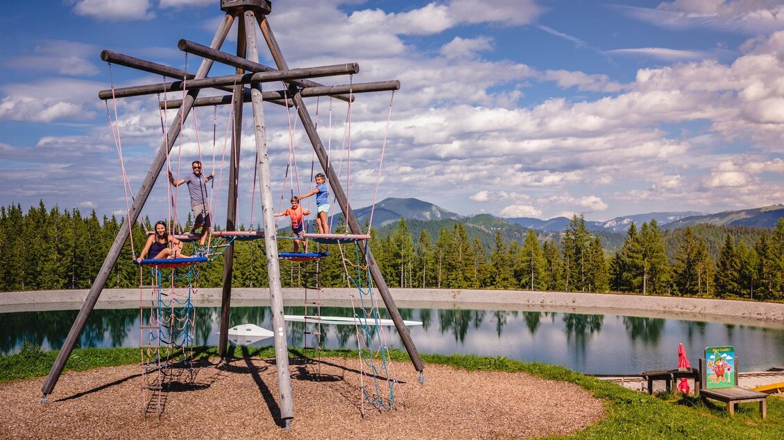 Ein großer Holzspielplatz mit einer Wippe und mehreren Kindern, die Spaß haben. Im Hintergrund sind Bäume und ein ruhiger See unter einem blauen Himmel zu sehen. | © Mariazeller Bürgeralpe
