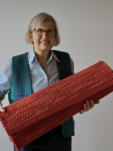 Eine freundliche Frau mit Brille hält einen roten Teppich. Sie trägt eine blaue Bluse und eine grüne Westenjacke. | © Strunz