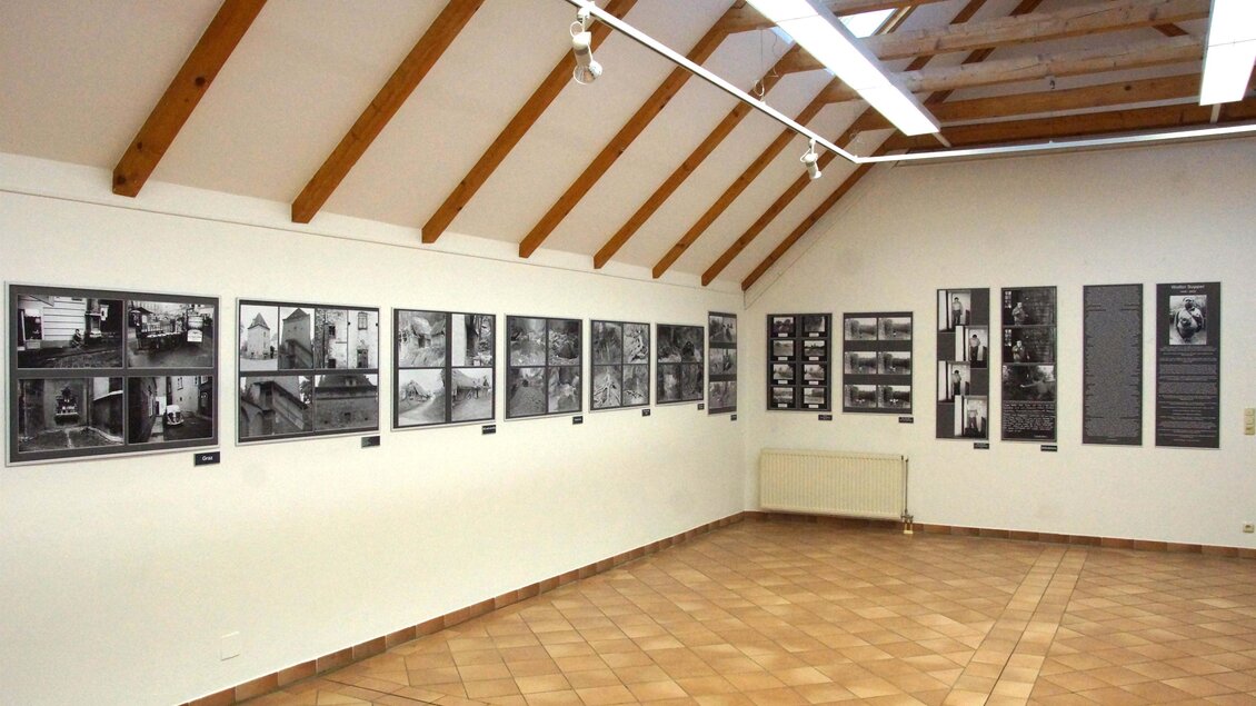Ein heller Ausstellungsraum mit Holzbalkendecke. An den Wänden hängen mehrere Schwarz-Weiß-Fotografien.